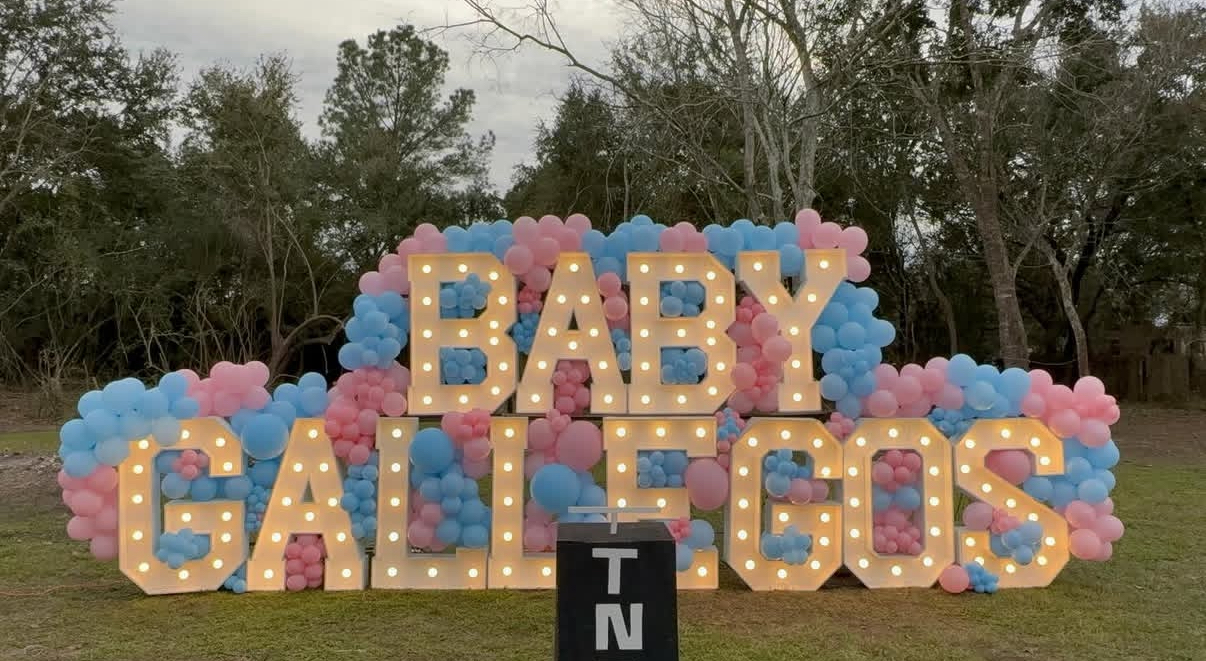 TNT gender reveal setup with BABY marquee letters, pink and blue balloon garland and outdoor trees